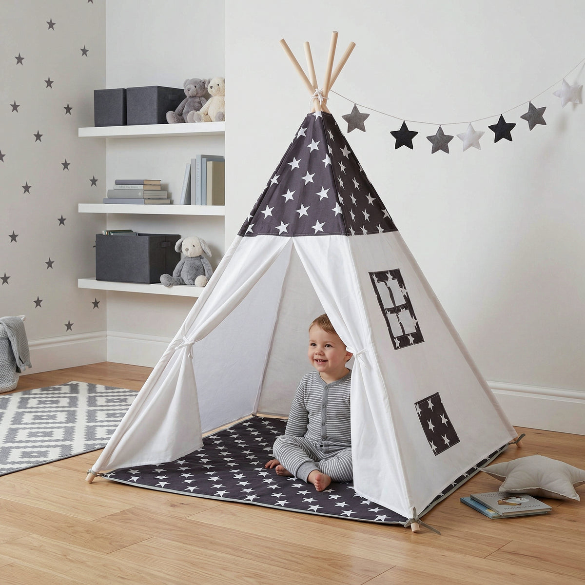 Niño pequeño sentado dentro de una tipi blanca y gris con diseño de estrellas en una recámara decorada con repisas blancas, peluches y alfombra geométrica - Tufan